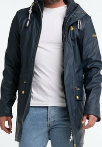 Veste imperméable bleu marine avec fermeture éclair à l'avant, capuche réglable, cordons blancs, accents jaunes et deux poches avant. Matière brillante et design mi-long.