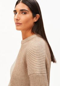 Beige gebreide sweater met een geribbelde textuur op de schouders, met een ronde hals en een zachte stofafwerking, gedragen door een model van opzij.