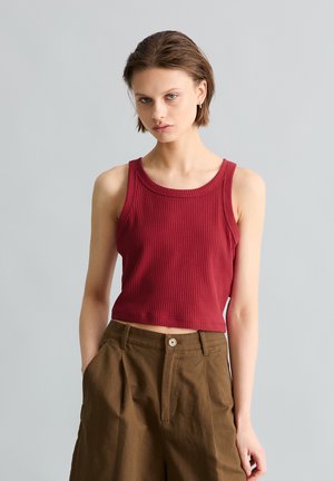 Jeune personne portant un crop top rouge côtelé sans manches et un pantalon marron taille haute, debout avec une main dans la poche sur un fond uni.