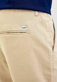 Šviesiai rudos kelnės su lygiu paviršiumi, su užpakaline kišenėle, ant kurios yra Jack & Jones etiketė ir siūti akcentai.