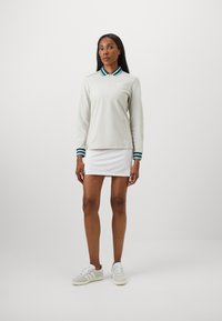 Vrouw staat in een lichtgrijs polo met lange mouwen, gestreepte kraag en manchet, witte rok en grijs-witte sneakers tegen een effen achtergrond.