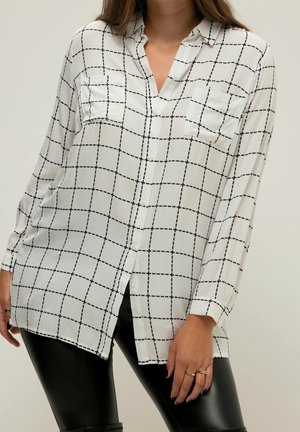 Femme portant une chemise blanche à manches longues avec un motif à carreaux noirs et deux poches poitrine, associée à un pantalon en cuir noir.