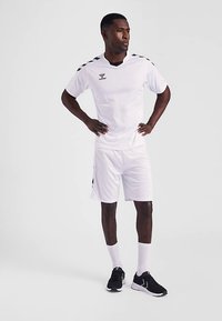 Weißes Sporttrikot und Shorts, schwarze Akzente an Schultern und Seiten. Dazu schwarze Sportschuhe und weiße Socken getragen.