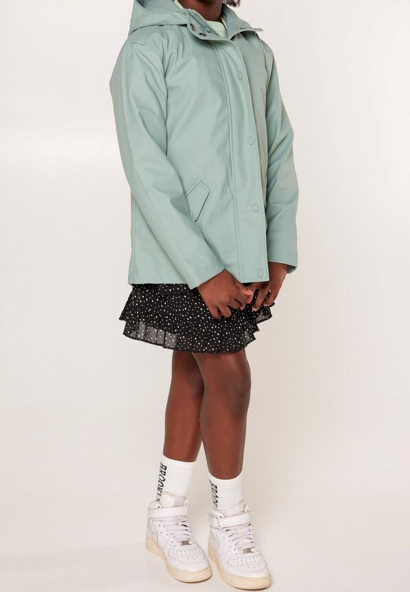 Light blue waterproof jacket with a hood, snap buttons, side pockets, layered over a black polka dot skirt, white sneakers, and crew socks.