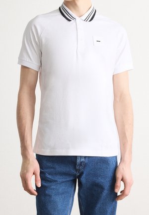 Mann trägt weißes Polohemd mit schwarzem Streifenkragen und kleinem „BOSS“-Logo auf der Brust, kombiniert mit blauen Jeans.