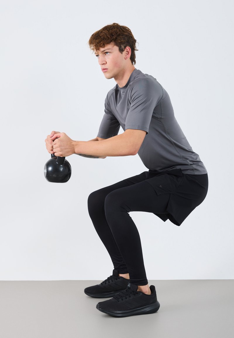 Fekete kettlebell, amelyet egy szürke sportpólót viselő és fekete leggingsben lévő személy tart, guggoló helyzetben, világosszürke padlón.