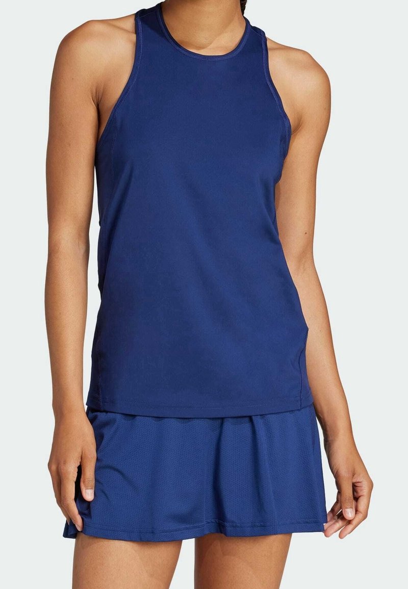 Femme portant un débardeur sportif bleu marine sans manches et une jupe de sport assortie, debout devant un fond clair uni.