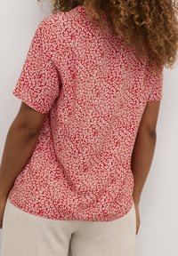 Krátký rukáv top v červené tkanině s malým bílým květinovým vzorem. Má uvolněný střih a zaoblený lem.