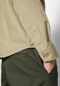 Camisa de pana color beige claro con puños abotonados, combinada con pantalones oscuros de corte ajustado en verde. Textura notable gracias a las estrías del tejido.