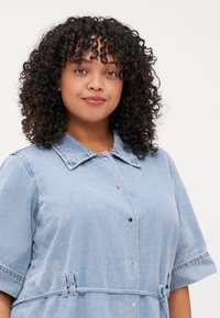 Lys blå denimkjole med korte ermer, stor krage og knapplukking foran. Har et knytbelte og subtil tekstur.