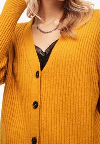 Senapsgul, ribbad cardigan med V-ringning, med svarta knappar och intrikata svarta spetsdetaljer under. Strukturerad stickad design.