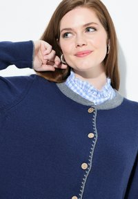 Vrouw met lichte huid en bruin haar in een blauwe knooptrui over een gerimpeld blauw-wit geblokt overhemd, lachend en haar haar aanrakend.
