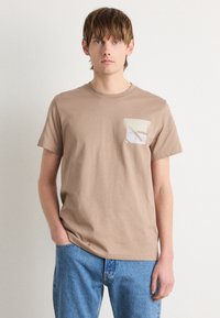 T-shirt en coton beige à manches courtes et col rond, avec une poche de couleur claire ornée d'un design géométrique. Pantalon en denim bleu.