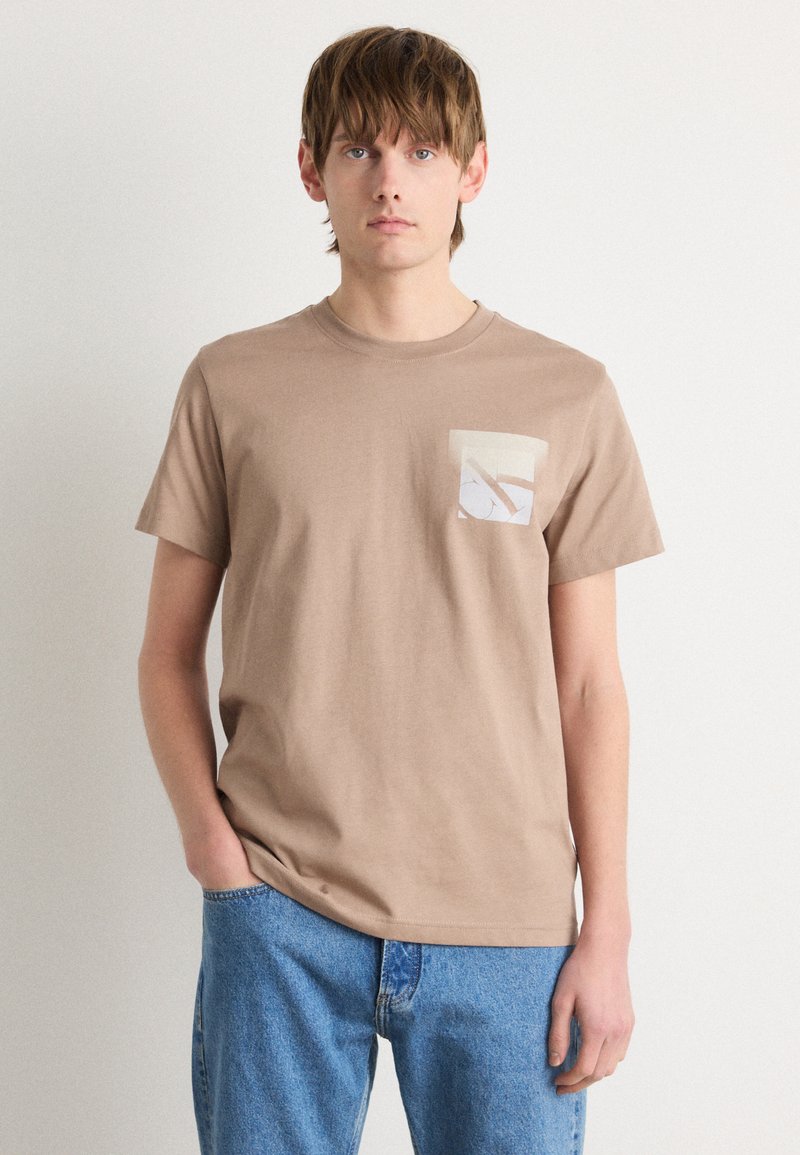 T-shirt en coton beige à manches courtes et col rond, avec une poche de couleur claire ornée d'un design géométrique. Pantalon en denim bleu.
