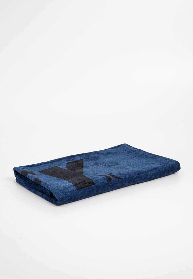 Serviette bleue, de forme rectangulaire, texture douce, présentant un grand motif graphique noir d'un côté. Pliée soigneusement sur un fond uni.