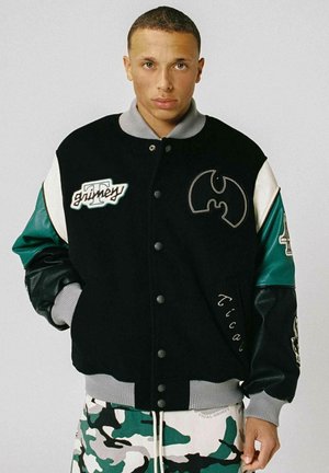 Joven con una chaqueta varsity negra, verde y blanca con parches y pantalones de camuflaje, de pie frente a un fondo liso.