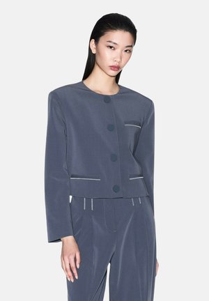 Giacca corta grigia con colletto rotondo, cinque grandi bottoni, tasche frontali con cuciture bianche a contrasto, abbinata a pantaloni grigi su misura.