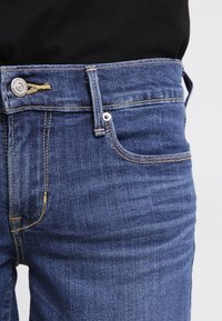 Mörkblå denimjeans med klassisk passform, femficksdesign, kontrasterande gula sömmar och silverfärgad knappstängning.