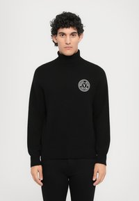 Μαύρο πλεκτό πουλόβερ με ψηλό γιακά, με κυκλικό λογότυπο σε λευκό και κείμενο "Versace Jeans Couture" στο στήθος. Ελαστικές ριμπ μανσέτες.