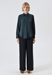 Dunkelgrünes Langarmhemd mit zwei Brusttaschen, geschwungener Saum, kombiniert mit weiten schwarzen Hosen und goldenen Schuhen mit Schleifenakzenten.