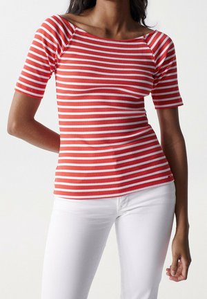 Vrouw draagt een rood-wit horizontaal gestreept topje met korte mouwen en een witte broek, staand voor een effen witte achtergrond.