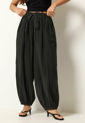 Vrouw draagt losse zwarte broek met trekkoord en geplooide enkels, gecombineerd met zwarte open-toe sandalen met hak en een zwarte cropped top.