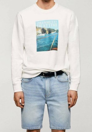 Persona con sudadera blanca con estampado de barco y mar, y shorts de mezclilla azul claro con cinturón negro, de pie contra un fondo liso.