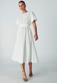 Biała, do kolan sukienka z bufkami i elastycznym pasem. Posiada prosty krój, gładką teksturę i lekką tkaninę. Czarne sandały na obcasie z paskami.