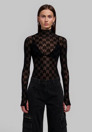 Mujer con el cabello castaño liso lleva una blusa negra semitransparente de manga larga con patrón repetido "W", bralette negro y pantalones cargo negros.