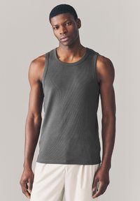 Débardeur gris côtelé avec un col rond et des emmanchures larges, porté par un mannequin masculin sur un fond neutre.