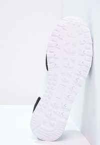 Bílá teniska s texturovanou gumovou podrážkou, se černým akcentem a hladkým svrchním materiálem. Zobrazuje design podrážky a vzory pro lepší grip.