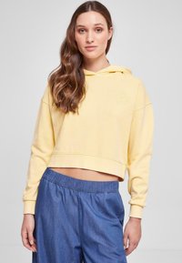 Felpa corta gialla con cappuccio regolabile e tasca frontale; presenta un tessuto texturizzato. Abbinata a pantaloni di jeans blu dal taglio largo.