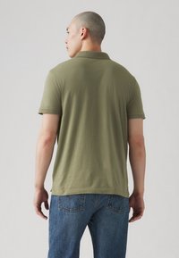 Polo din bumbac de culoare verde măsliniu, cu mâneci scurte, fante laterale și guler clasic, purtat cu blugi din denim albastru.