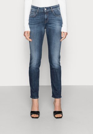 Femme portant un jean bleu slim à taille mi-haute avec un haut blanc côtelé à manches longues et des sandales noires à talons ouvertes à l\'avant.