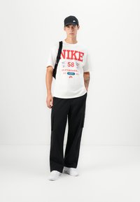 White T-shirt with red Nike graphic, black cargo pants, and white sneakers. Black cap with Nike logo. Simple, sporty design and casual fit.