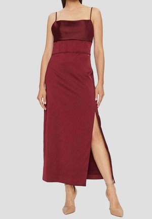 Femme portant une robe midi bordeaux sans manches avec une fente latérale et des talons nude à bride cheville, debout devant un fond uni.