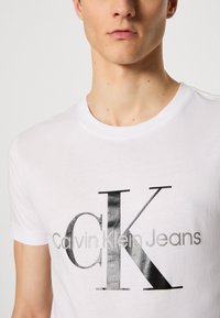 Balta kokvilnas T-kreklu ar melnu metalisku logo uzdrukājumu, kas nolasāms "CK Calvin Klein Jeans". Īsas piedurknes un klasiskā apaļā kakla dizains.