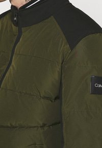 Giacca puffer verde oliva con accenti neri, chiusura con zip, colletto alto, texture trapuntata e una toppa con logo sulla manica sinistra.