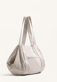 Beige tygväska med rundad form, dubbla platta handtag och en framficka. Jämn textur med subtila sömdetaljer.