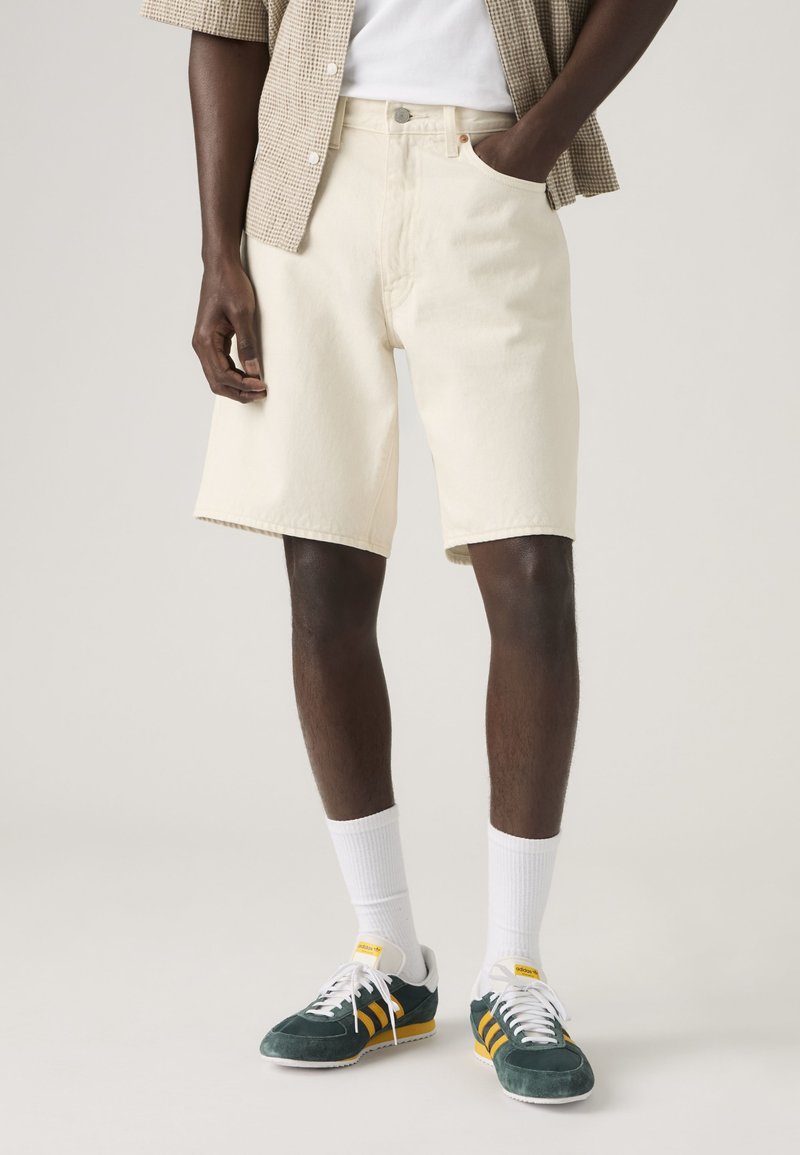 Uomo con pantaloncini color crema fino al ginocchio, calzini bianchi e scarpe da ginnastica verdi con strisce gialle, in piedi davanti a uno sfondo uniforme.