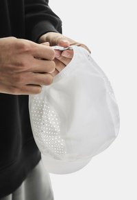 Witte pet van ademend meshmateriaal, met kleine perforaties voor ventilatie. Een verstelbare band is zichtbaar. Handen houden de pet vast.