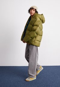 Zelená péřová bunda s kapucí, zapínáním na zip a prošívaným designem. V kombinaci s šedými kalhotami širokého střihu a šedými botami s žlutými akcenty.