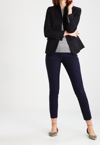 Blazer negro ajustado con cuello alto, top gris entallado y pantalones slim-fit en color azul marino. El conjunto se completa con bailarinas plateadas.