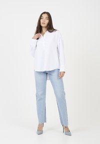 Donna che indossa una camicia bianca larga con bottoni, jeans dritti azzurri chiari e scarpe con tacco grigie, in piedi di fronte a uno sfondo bianco semplice.