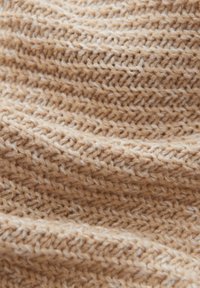 Tissu tricoté beige avec un motif texturé, présentant un mélange de fibres claires et foncées, créant une apparence douce et chaleureuse.