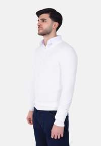 Maglione bianco a scollo a V realizzato in un tessuto morbido e leggero. Presenta maniche lunghe, polsini e orlo a costine. Abbinato a jeans blu scuro.