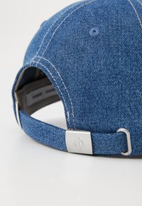 Gorra de mezclilla con una corona estructurada, tela azul, costuras contrastantes y un cierre de hebilla metálica en la parte posterior con un detalle de logo.