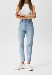 Jeans de tiro alto azul claro con rasgaduras en las rodillas, combinados con una camiseta blanca sin mangas ajustada y zapatillas blancas, de pie sobre un fondo liso.