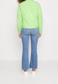 Grön denimjacka med knäppningar, kombinerad med ljusblå flare-jeans och vita sneakers, sedd bakifrån.