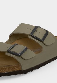 Närbild på beige sandal med två justerbara svarta metallspännen och korkfotsula på vit bakgrund.